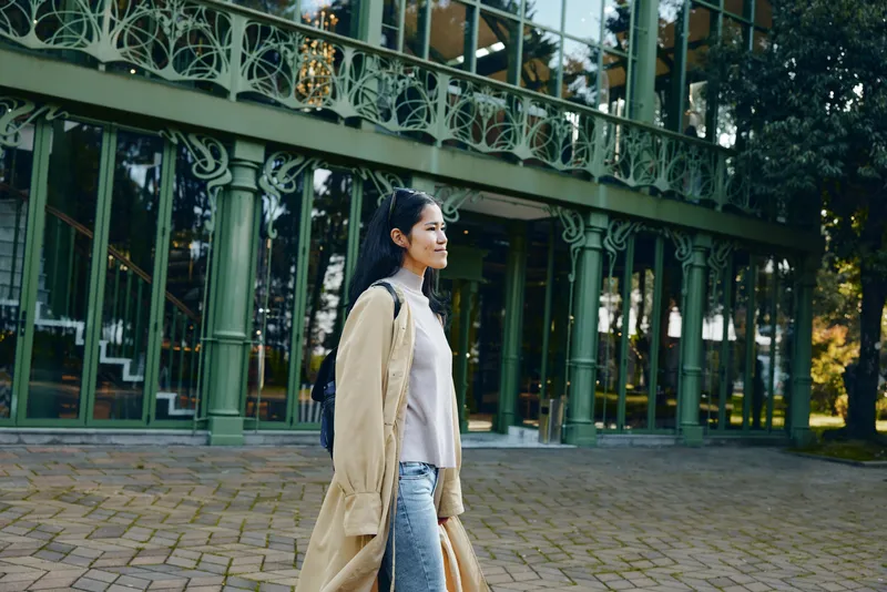 Woman, coat, outdoor, fashion, urban, street, model, confident, chic, style, strolls beside a green iron railing in a park setting, blending architectural detail with casual elegance and natural