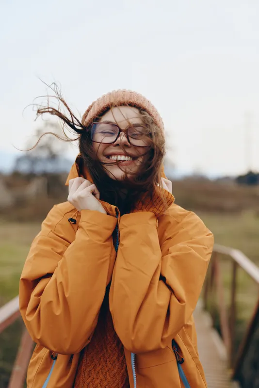 Smiling woman in winter coat and scarf outdoors, wearing glasses, hair lifted by wind. She laughs on a sunny day in a countryside field, lifestyle moment.