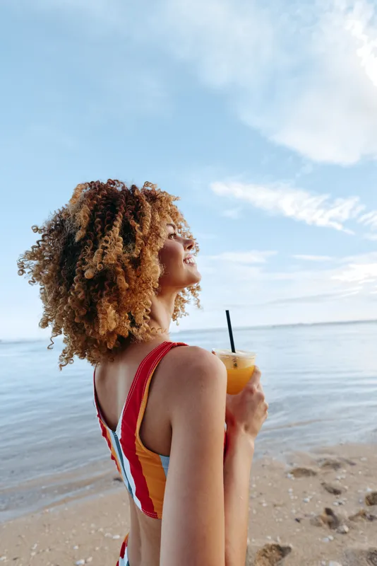 Young woman with curly hair enjoys a refreshing drink while standing on a sandy beach, looking up at the clear sky with a relaxed and happy expression.