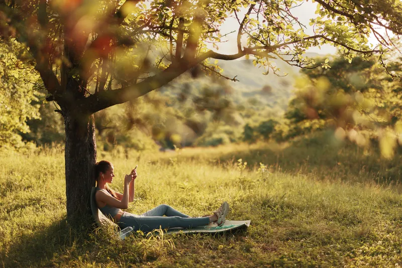 Young woman enjoying nature while using her smartphone under a tree in a sunny field, dressed casually in comfortable attire, representing a peaceful lifestyle