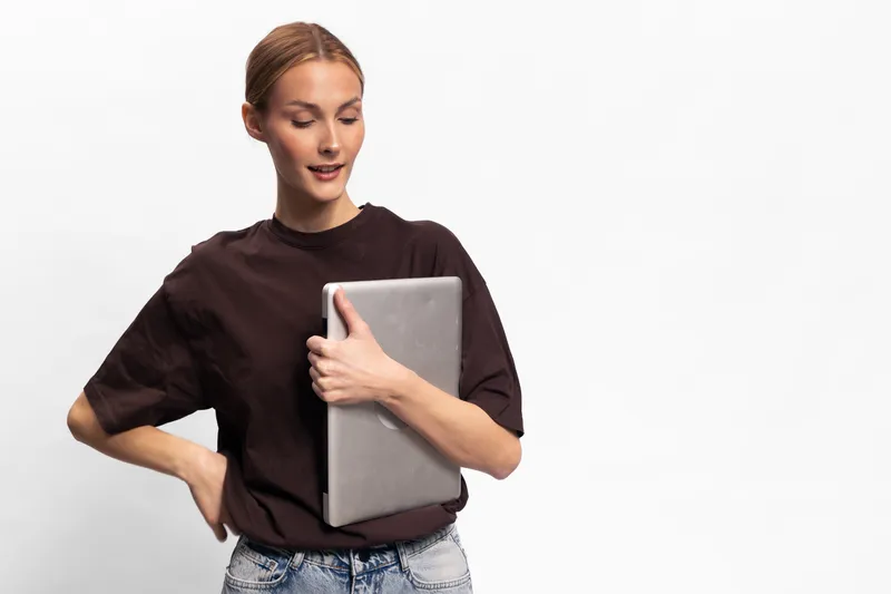 Young woman in casual clothes holds laptop with a slight smile, standing against white background. Concept for technology, remote work, and casual lifestyle with digital device.
