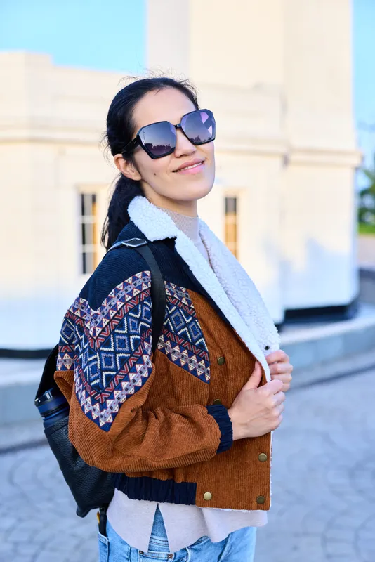 Fashion, style, jacket, sunglasses, backpack, urban, confident, outdoor scene with a woman wearing a patterned jacket and scarf, smiling under bright daylight, showcasing casual chic and modern vibe