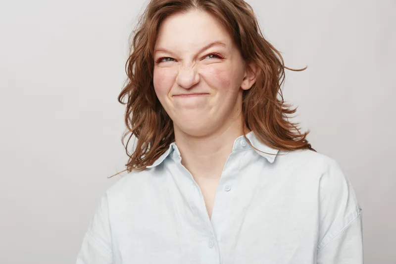 Young woman with wavy brown hair makes a funny grimace wearing a white shirt on light background. Concept of emotions, humor, and playful facial expression in casual style.