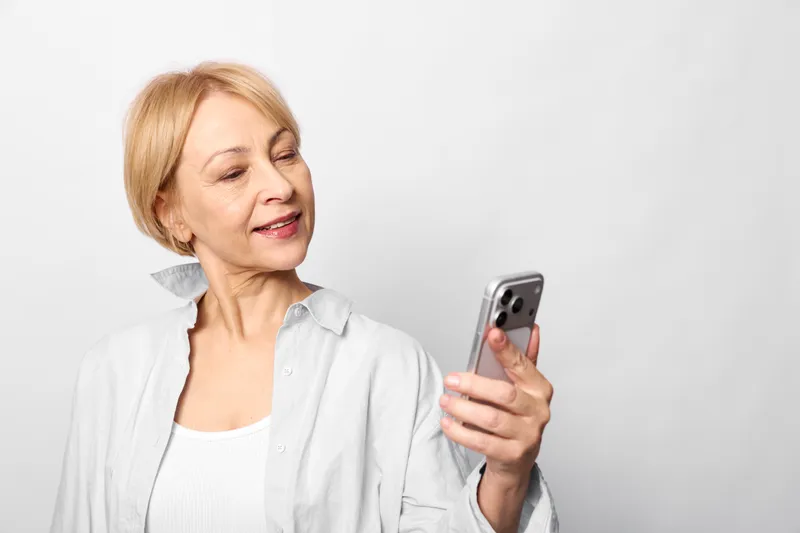 Elderly woman portrait smiling indoors, holding smartphone for communication and messaging, using technology for texting, casual white clothing, neutral background
