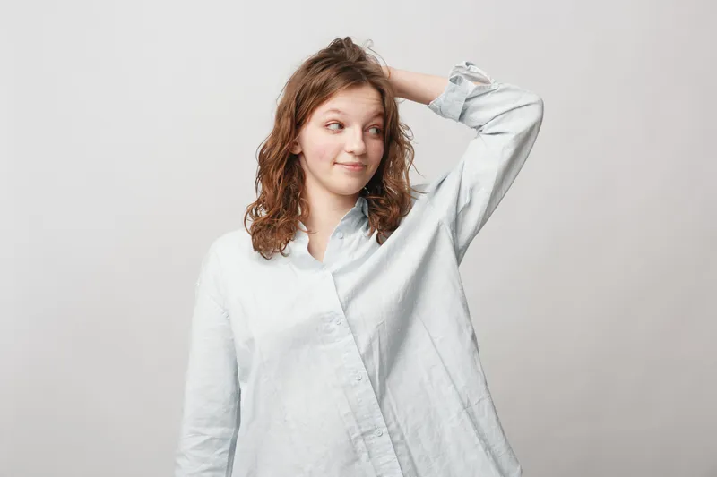 Young woman with curly hair in white shirt looks sideways with slight smile and hand behind head. Casual portrait on light background. Concept of hesitation, thoughtfulness, and uncertainty.