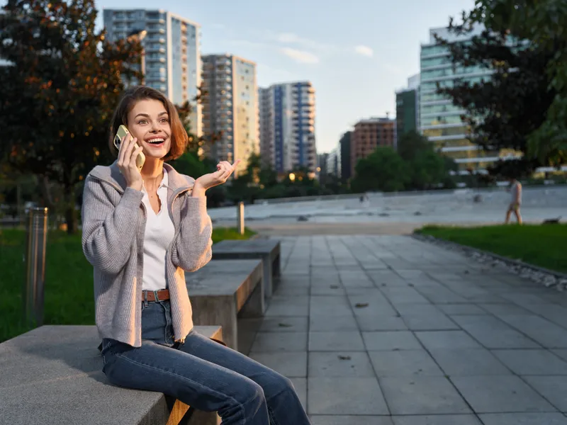 A cheerful young woman sits on a park bench in an urban plaza, talking on a smartphone. She smiles brightly, enjoying a lively moment in a modern city setting.