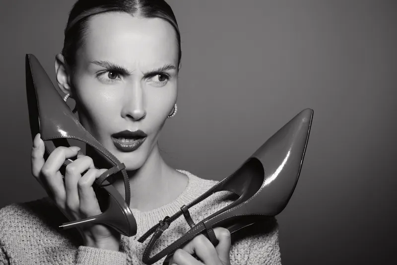 Caucasian woman with light skin looks confused holding red high heel shoes near face on grey background. Concept for fashion, footwear, and style.