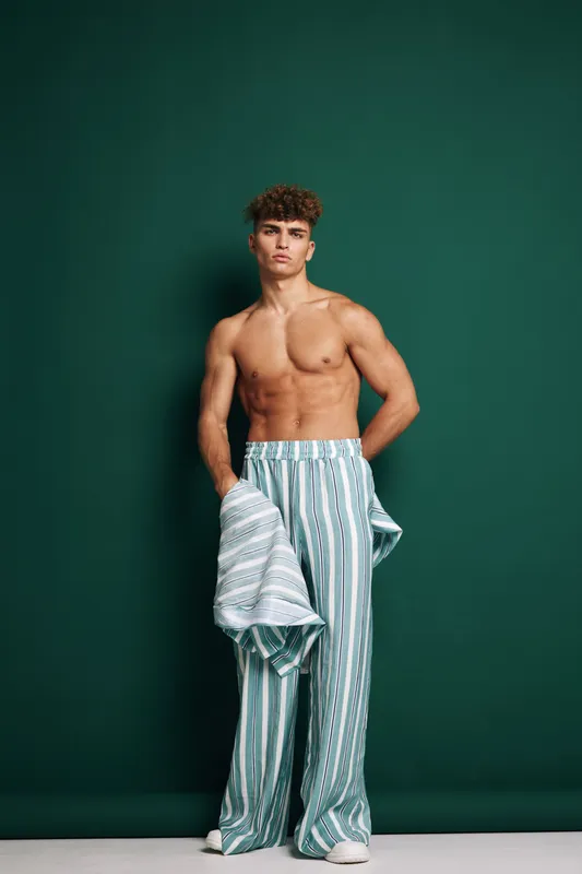 Young man with curly hair posing shirtless wearing light colored striped loose pants holding a modern jacket. Fashion portrait with confident model on green background in studio.