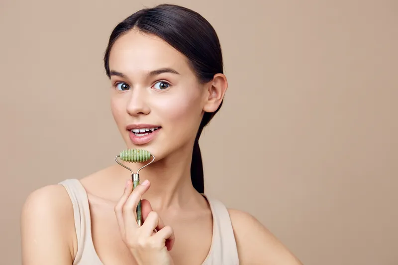 Young woman smiling with a jade facial roller, promoting skincare and wellness, neutral background highlighting natural beauty and self care routines