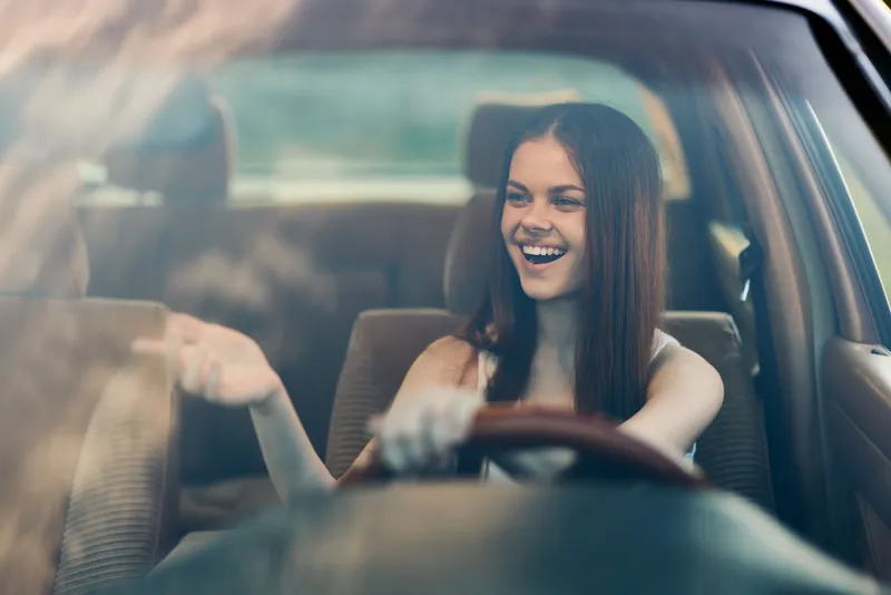 Young woman joyfully driving a car, exuding happiness and freedom, with sunlight streaming through the windows, showcasing a vibrant and cheerful mood.