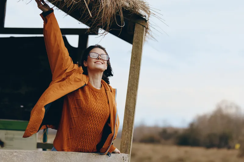 Woman lifestyle outdoors in spring sunshine, orange sweater and sunglasses, smiling while resting beside a wooden post, arms raised, calm mood, natural landscape background soft light