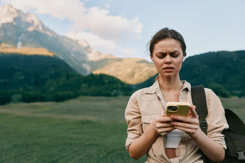 Young woman outdoors using smartphone with focused expression, casual outfit, natural mountain landscape background, daytime, lifestyle concept