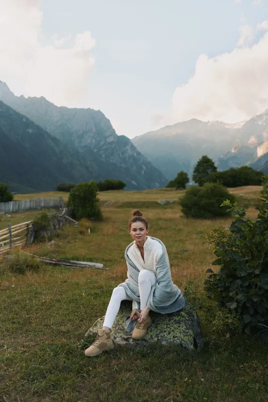 Casual woman sitting on a rock in an open field with distant mountains, showing a relaxed outdoor lifestyle and connection with nature under soft daylight.