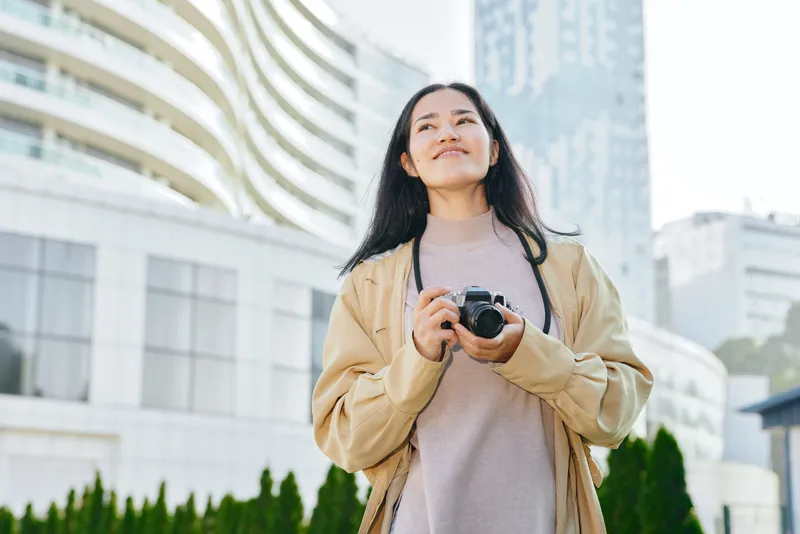 Camera in hand, woman gazes at a modern cityscape, smiling softly, wearing a beige jacket and knit sweater, captured outdoors among tall buildings and green shrubbery