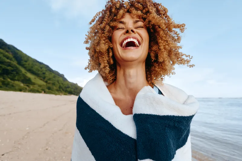 Happy woman with curly hair wrapped in a towel laughing on the beach near the ocean. Sunny day with blue sky and green hills in the background. Concept of summer and joy.
