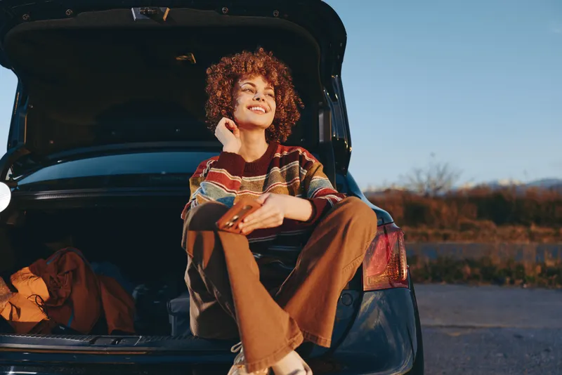 Woman in casual jacket sits in car trunk during road trip, smiling and laughing outdoors at sunset, holding luggage in open parked car, lifestyle portrait, autumn countryside.