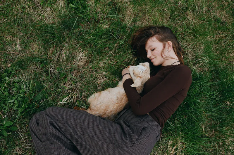 Relaxed woman lying on grass, cuddling a small pet, enjoying a peaceful outdoor moment, wearing a warm casual outfit, intimate mood, natural light, lifestyle scene conveying affectionate bond in a