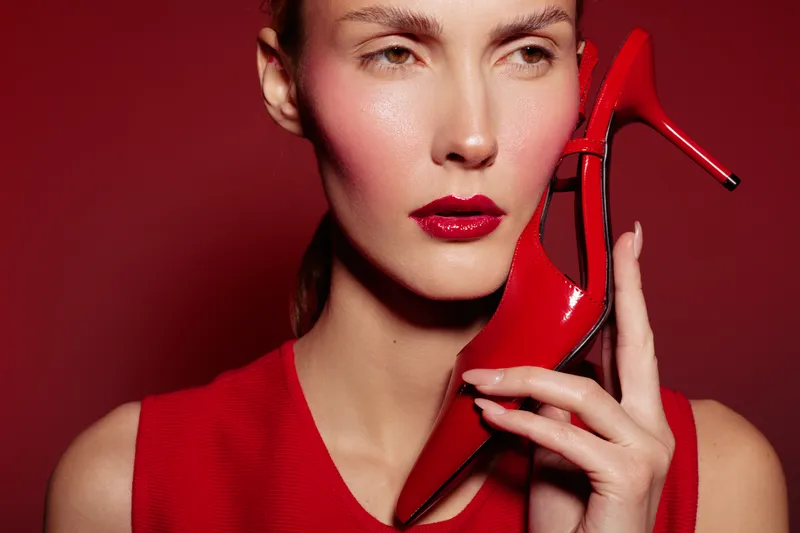 Close-up of a stylish woman with red lipstick holding a red high heel shoe near her face. Fashion concept with bold makeup and matching red outfit on red background.
