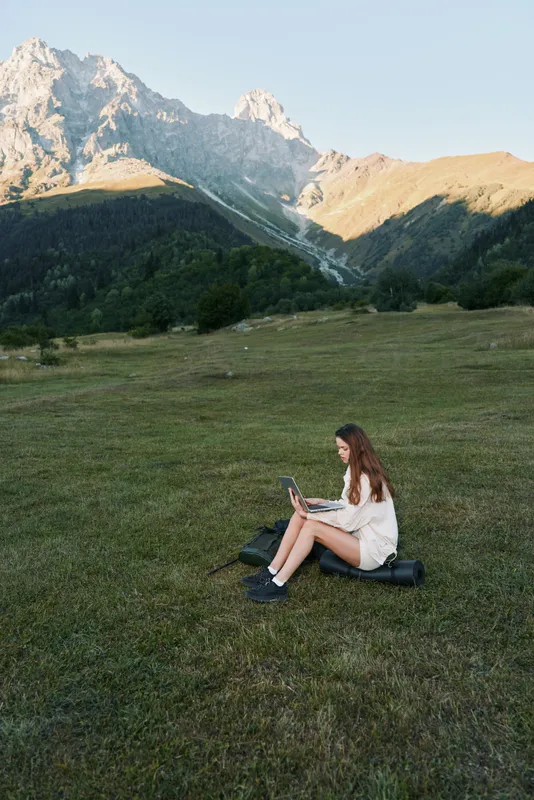 A woman sits on grassy terrain in a scenic mountain landscape, using a tablet outdoors. She appears focused and calm, creating a peaceful and productive moment in nature.