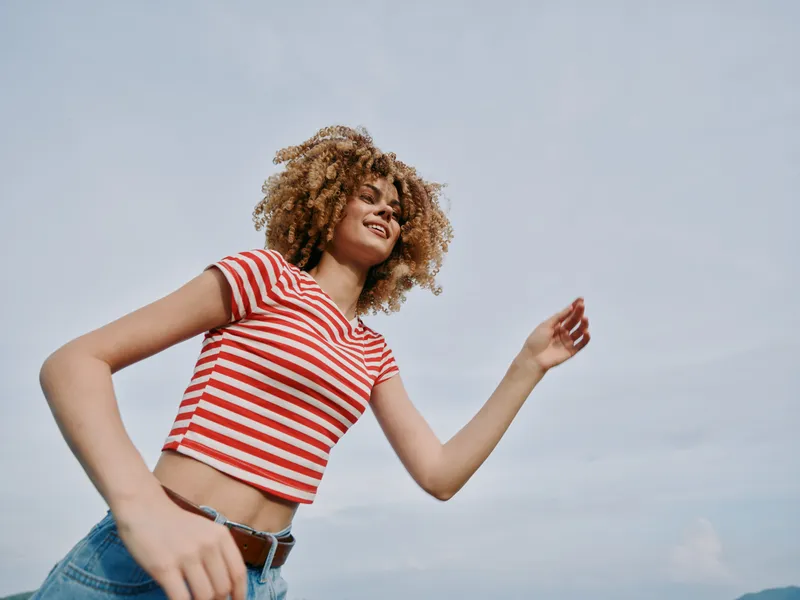 Dynamic portrait of a young woman with curly hair wearing a red striped crop top and denim jeans, captured mid stride outdoors against a bright, open sky.