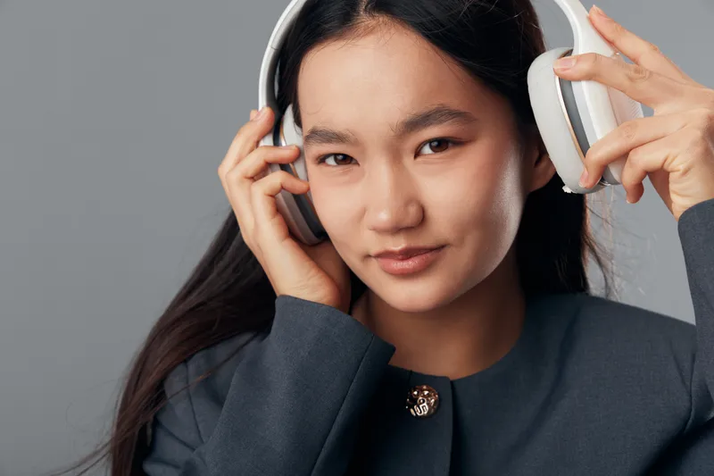 Young woman wearing large headphones, adjusting them with hands, looking at camera. Portrait with straight dark hair and neutral background, casual clothing with detailed buttons.