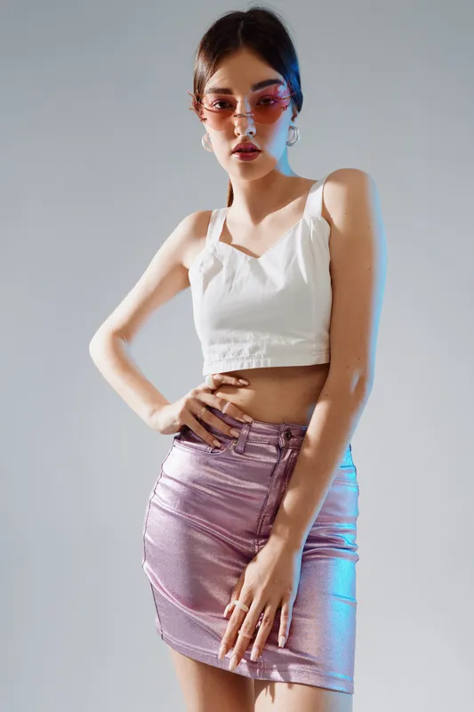 Young woman in a white crop top and metallic pink skirt poses confidently in studio lighting. She wears stylish sunglasses and hoop earrings. Concept of beauty, fashion, and style.