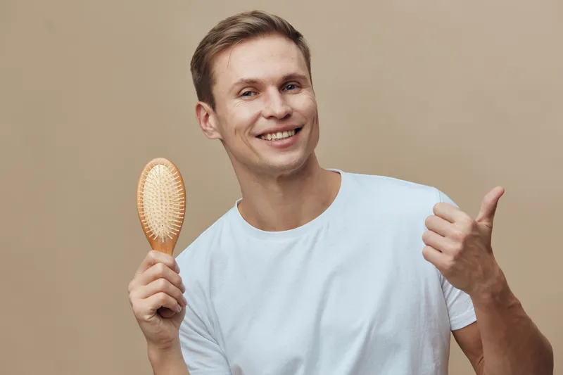 Smiling man with short brown hair holding a wooden hairbrush, showing thumbs up gesture, casual white t-shirt, isolated on beige background, studio portrait, positive emotion, lifestyle concept