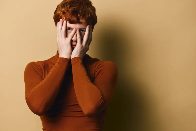 Young man with red hair wearing orange turtleneck covering face with hands isolated on beige background. Portrait of male model expressing emotion or shyness in studio.