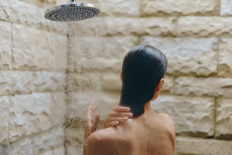 Woman in outdoor shower washing hair with water falling down against stone wall background in natural light setting, fresh and relaxing moment.