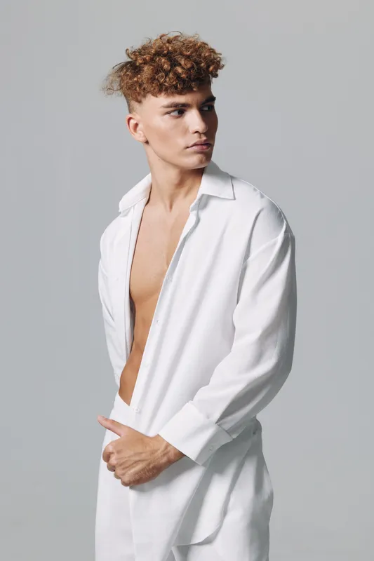 Young man with curly hair wearing white open shirt and pants posing against gray background in studio. Fashion model with serious expression and casual style.