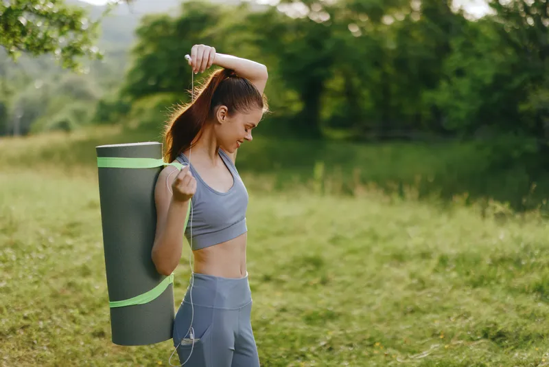 Young athletic woman with yoga mat outdoors, smiling in activewear, enjoying nature and fitness in a sunny, green environment