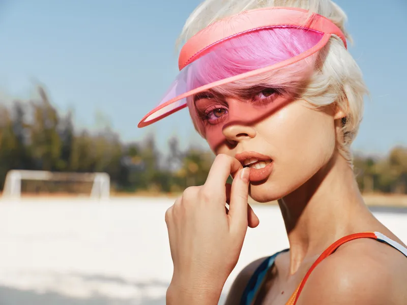 Close up portrait of a stylish person on a sunny beach wearing a pink visor, pale hair, and sunlit skin, conveying bold summer fashion and playful confidence.