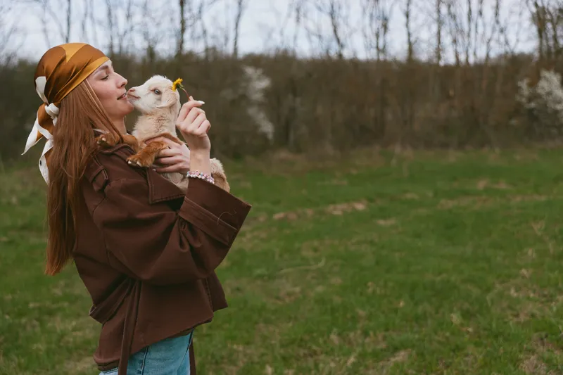 Young woman with long red hair holds a small goat in her arms while standing on a grassy field in autumn wearing a brown jacket and orange headscarf.