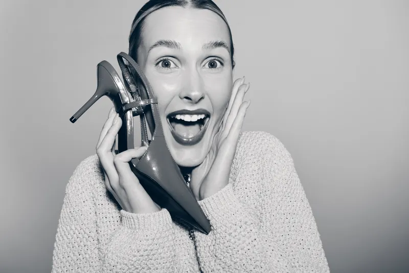 Excited white woman with fair skin holds shiny high heel shoe near face, smiling widely with open mouth. Concept of fashion, joy, and style.