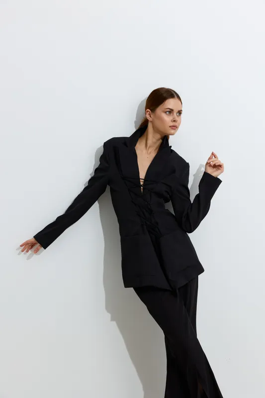 Fashion woman dressed in black stylish outfit posing against plain white wall, showcasing modern elegance and minimalistic beauty with confident posture and sleek hairstyle.