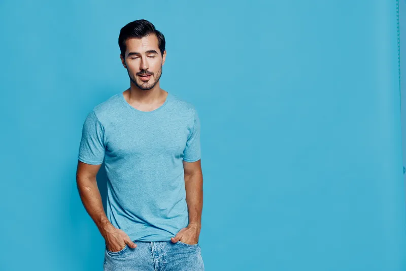 Smiling young caucasian man in a casual blue t-shirt stands against a bright blue background with a relaxed expression, hands in pockets. Lifestyle concept.