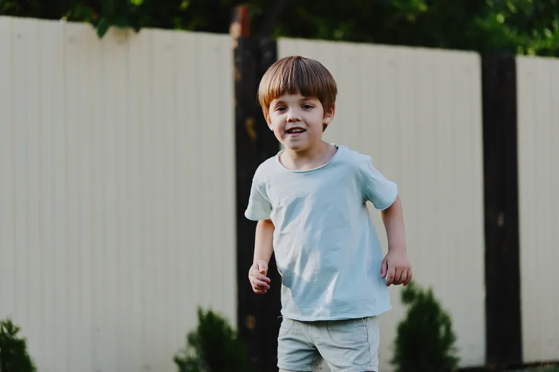 Active boy playing outdoors, wearing a light blue t-shirt and shorts, joyful expression amidst a green garden background, capturing the essence of childhood fun.