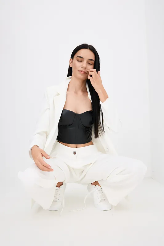 young woman in white suit and black top squatting with closed eyes, showcasing modern fashion against minimalistic white background, calm and poised expression