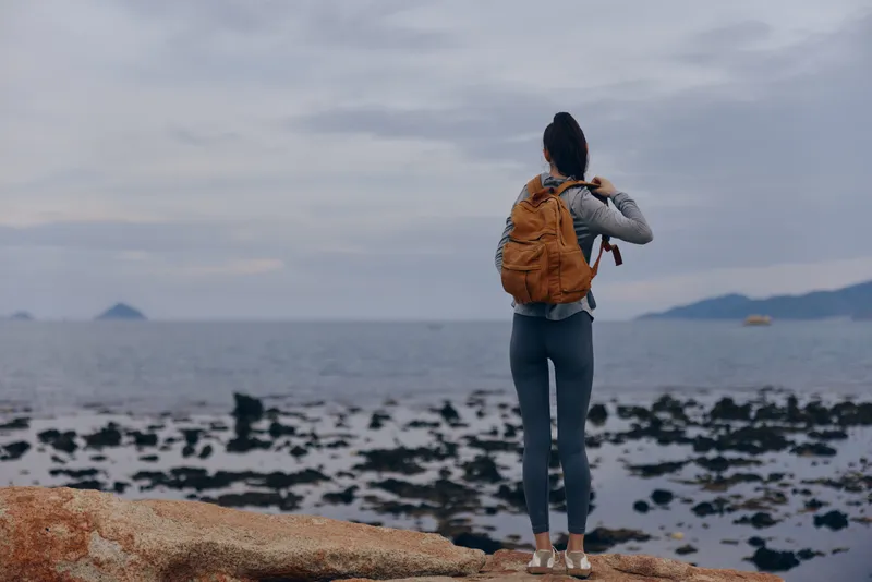Young woman with a backpack standing on rocky beach, gazing at ocean view, wearing casual athletic wear Adventure, travel, exploration