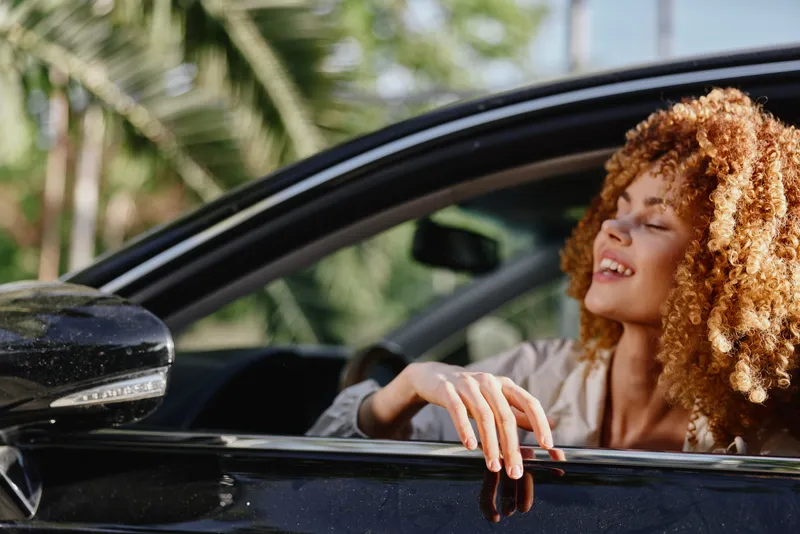 happy woman with curly hair enjoying summer drive in black car, smiling with eyes closed, tropical background, natural light, carefree mood, outdoor lifestyle