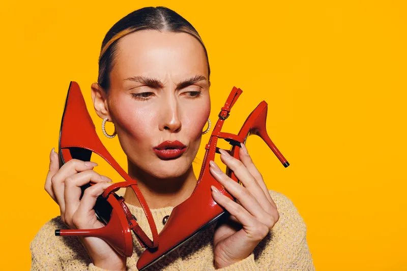 Young woman with slicked hair holds red high heels near face, making a confused expression on bright orange background. Concept for fashion, style, and footwear choice.