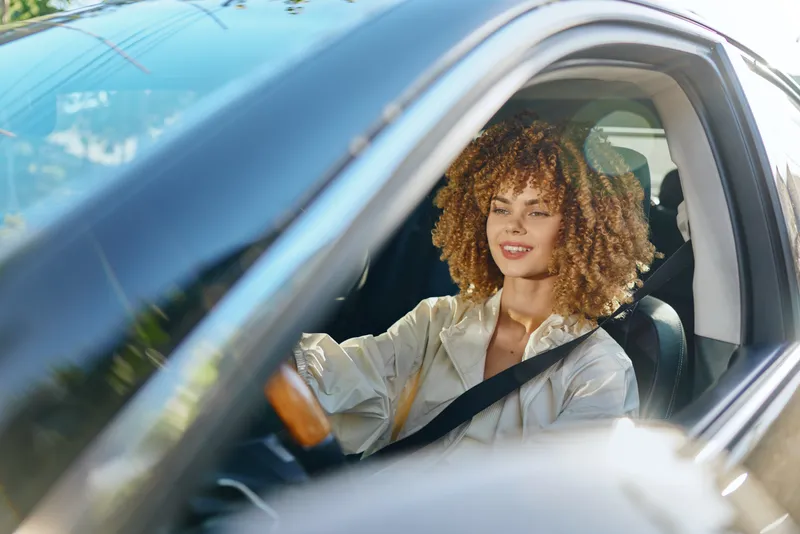 Smiling woman driving a car wearing seatbelt with curly hair in natural light, casual outfit, relaxed mood, modern interior, safety concept.