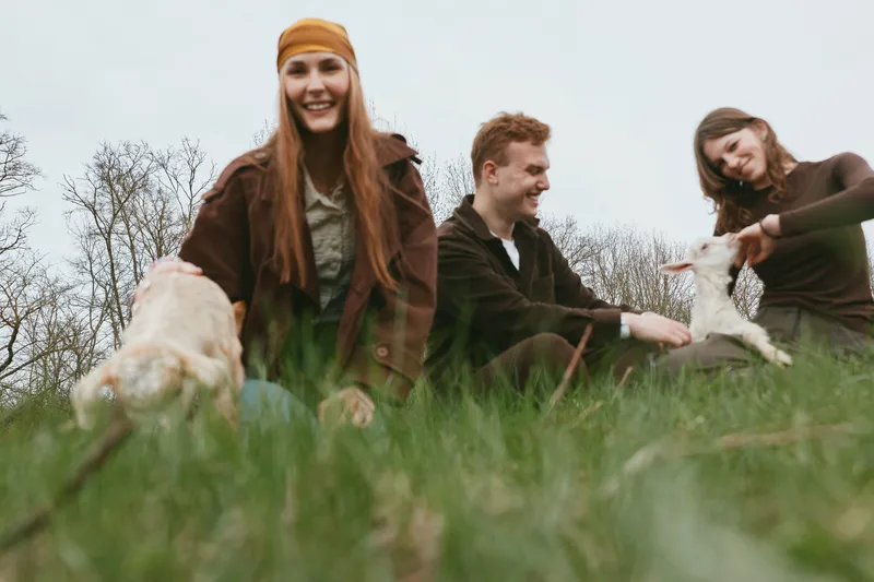Cheerful outdoor scene with friends and goats in a grassy field, candid laughter, relaxed posture, natural colors, soft breeze, sunlit moment, lifestyle vibe, kids nearby, animal companionship