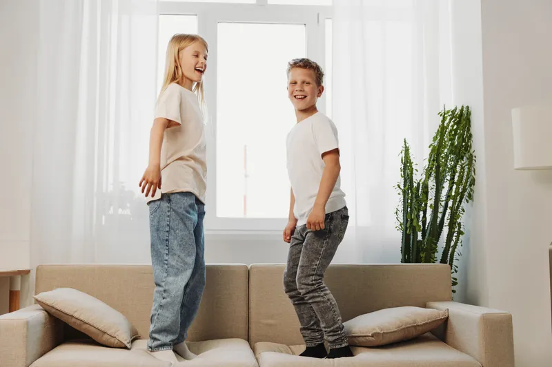 Two happy children jumping on a beige sofa in a bright living room, enjoying playful moment with smiles and casual clothing. Concept of fun and childhood joy.