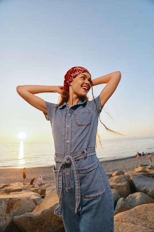 Lifestyle woman in modern boho-western denim street style enjoys sunset by the beach, smiling confidently with a red bandana and relaxed natural pose in warm film color tones.