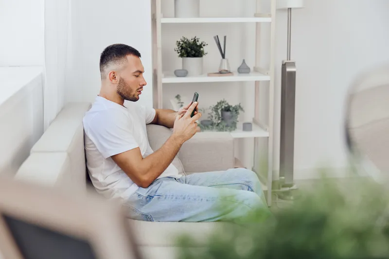 Engaged man using smartphone while relaxing on a cozy sofa, surrounded by modern decor and greenery, showcasing a serene and contemporary living space.