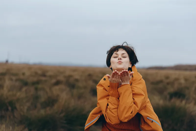 Child outdoor coat hands autumn wind overcast, girl in rural field holds coat sleeves near face, warm orange jacket, gentle smile, calm lifestyle, cloudy sky and grass background, very soft