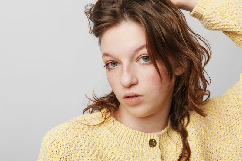 Young woman with natural skin and brown hair wearing yellow knitted sweater looks thoughtfully at camera on light background. Concept of natural beauty and casual fashion.