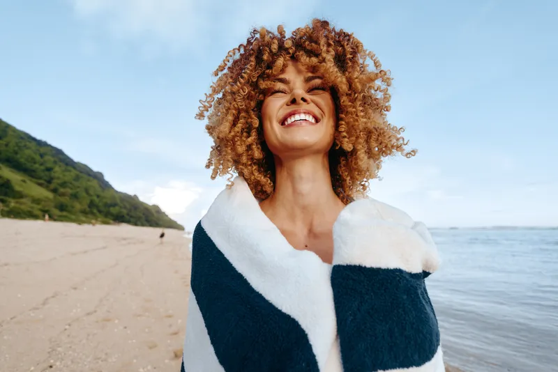 Happy young woman with curly hair wrapped in striped blanket smiles joyfully on beach near calm sea with blue sky and green hills in background