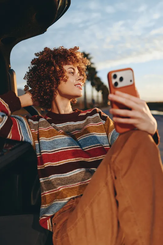 Woman selfie with smartphone while seated outdoors, curly hair framing face, striped sweater, relaxed lifestyle mood during golden hour, she looks at screen, blurred park background